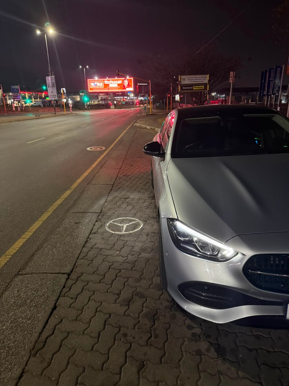 The front corner of the Mercedes at night beside a long empty road, badge projected on the tarmac